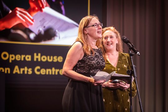 Two staff members on stage, one is stood at the microphone giving a speech