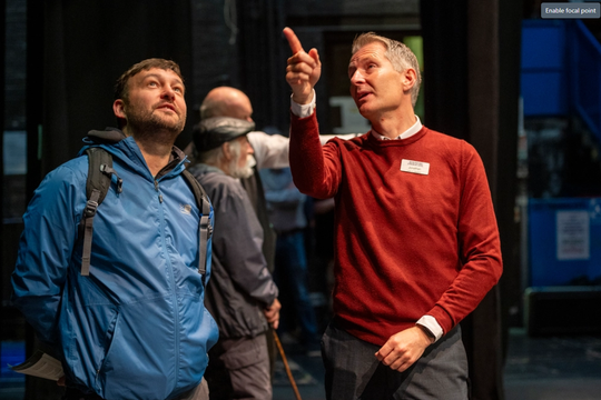 Staff member in red sweater points upwards while speaking to a man in a blue waterproof