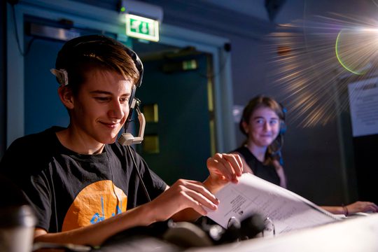 Two young people with headphones on running sound deck for a show, smiling and looking at script