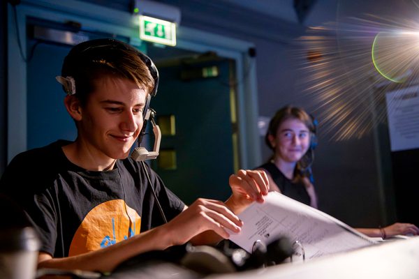 Two young people with headphones on running sound deck for a show, smiling and looking at script