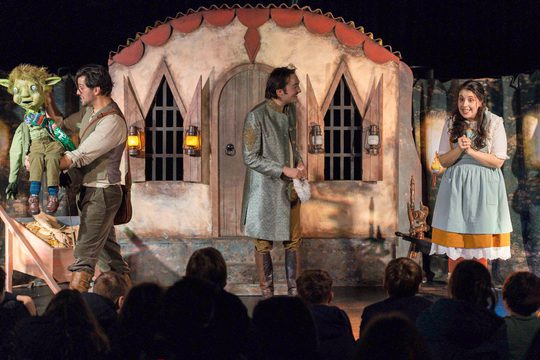 Three performers and puppet with house set in the background. At the forefront is the back of the heads of audience members