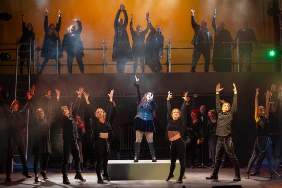 performers on stage, all dressed in black with hands in the air. Lead singer in centre stage on platform