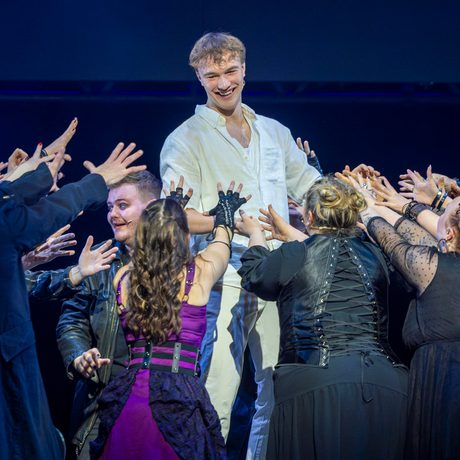 Cast on stage, group of people reaching towards one man dressed in white shirt