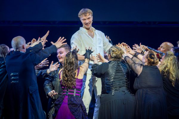 Cast on stage, group of people reaching towards one man dressed in white shirt