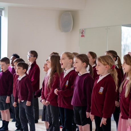 Image of school children in a red uniform singing in a rehearsal room with a wall of mirrors behind