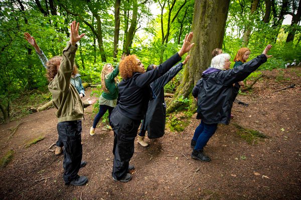 Group of adult performers in woodland, all with arms stretched to the sky
