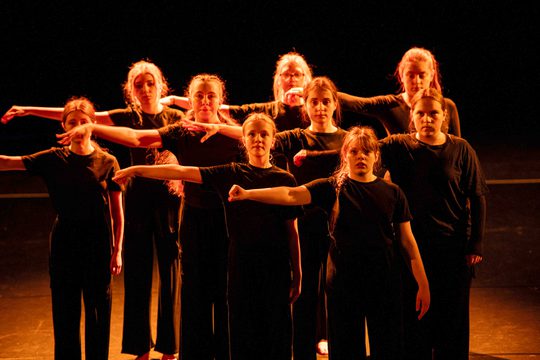 Dancers on stage, all dressed in black holding out left arm to the side