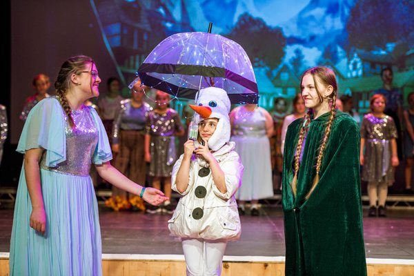 Three young people performing in Frozen Jr. musical, two in dresses and one in snowman outfit holding umbrella