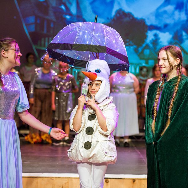 Three young people performing in Frozen Jr. musical, two in dresses and one in snowman outfit holding umbrella