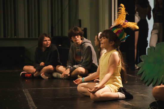 young performers with feather crown sits, with her hands on her knees, singing to the other children sat around the room