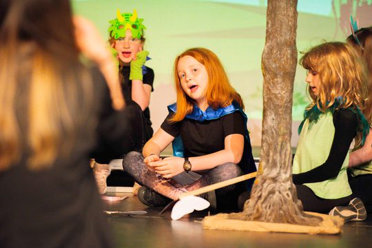 Child sat cross legged on stage looking at teacher at front of stage