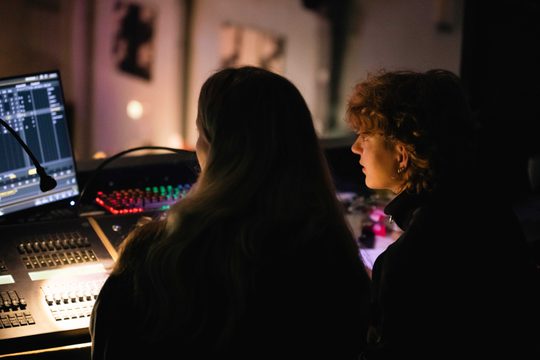 Two young people sat at sound and lighting desk, taken from behind