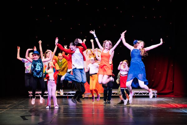 A group of young people wearing colourful costumes jump with their arms in the air. They are all smiling.