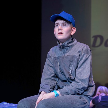 Young performer in cap and grey tracksuit, sat on bed looking into audience