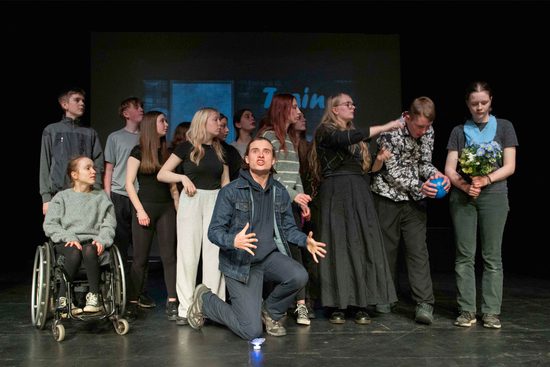 Group of young performers on stage, central performer on his knees looking into audience