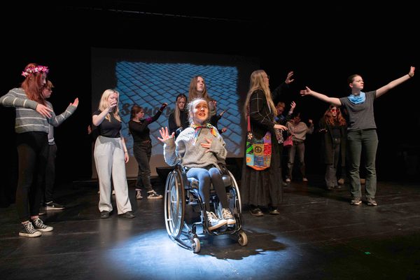 Group of young people performing on stage, performer in a wheelchair centre stage illuminated by spotlight