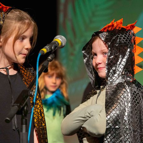 Two young performers stood at a microphone, their both wear metallic capes with dragon scales up the hoods