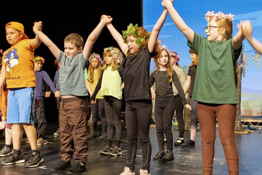 Young performers in a row holding hands in the air as they go to bow to audience.