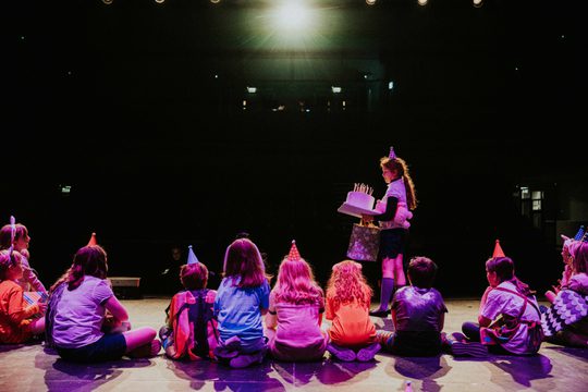 Group of young people sat on stage wearing party hats, one child standing holding birthday cake