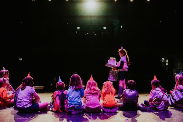 Group of young people sat on stage wearing party hats, one child standing holding birthday cake