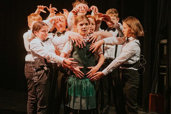 Group of young people on stage, all dressed in suits and bow ties with arms wrapped around young girl in centre