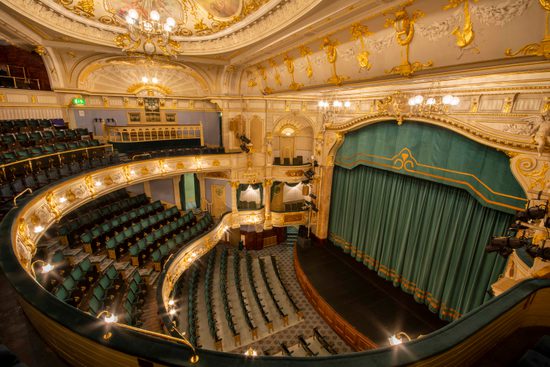 View of the inside of the opera house from the side of upper circle, the stage curtains are closed and match the green of the seats. Ornate gold plaster work covers the ceiling