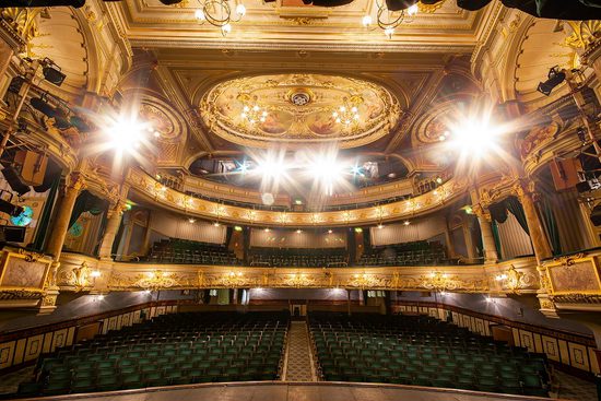interior of the opera house from the stage, the green seats of the stalls are visible through the stage lights, as is the ornate gold ceiling