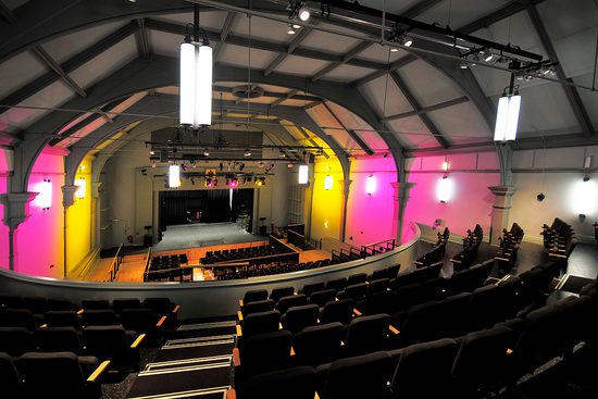 The pavilion art's centre taken from the back of the audience. Yellow and pink lights beside the stage.