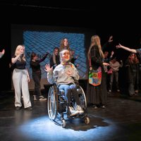Performers on stage with projection screen in back, in the centre a performer in a wheelchair in the spotlight