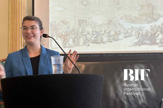 A woman with brunette hair, wearing a black top, a blue blazer and glasses talks into a microphone, cup of water in front of her, projector screen behind her showing a painting of people from the late eighteenth century.