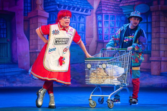 A panto dame in a red dress, white apron, and red wig leans on a shopping trolley.