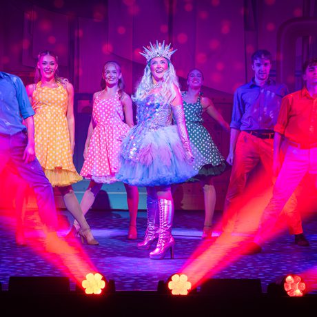 Various people in sparkly and colourful costumes smile. They are framed by red strobe lights.