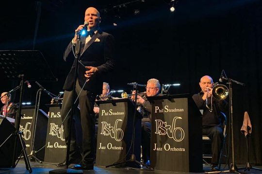 A man in a black suit and bow tie singing into a microphone, with instrumentalists sat behind him.