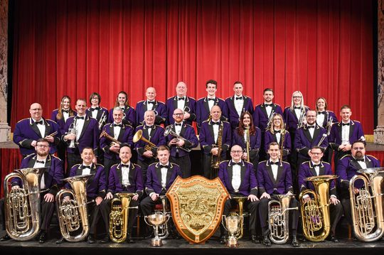 A brass band wearing purple jackets and holding their instruments standing around a large golden crest in front of a red curtain.