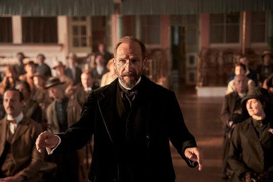 A man in a black suit and glasses stands conducting with his back to the audience.