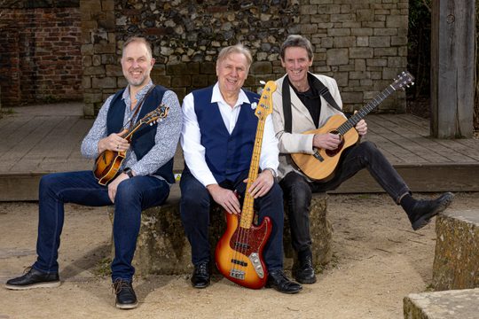 Three men holding guitars sitting on a stone.