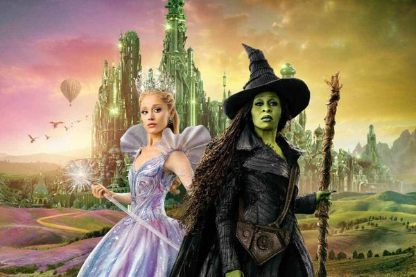 Elphaba, in a black dress and witch's hat, and Glinda, in a purple sparkly dress, stand in front of Oz.