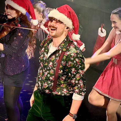 A man in green trousers, a green shirt with Santa on it, red suspenders and a Santa hat.