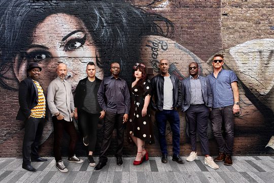 Eight members of the band stand in front of a street mural of Amy Winehouse