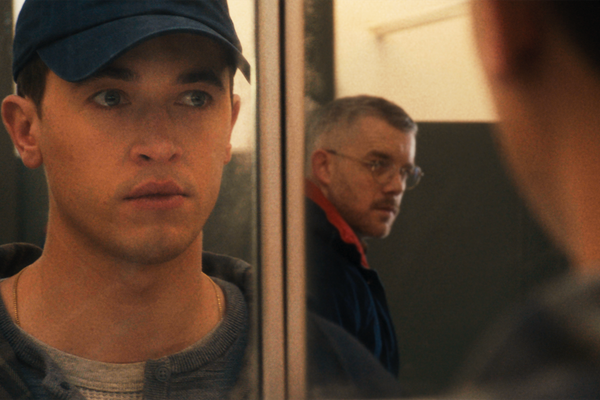 A man wearing a cap looks in the mirror and sees another man looking over his shoulder.