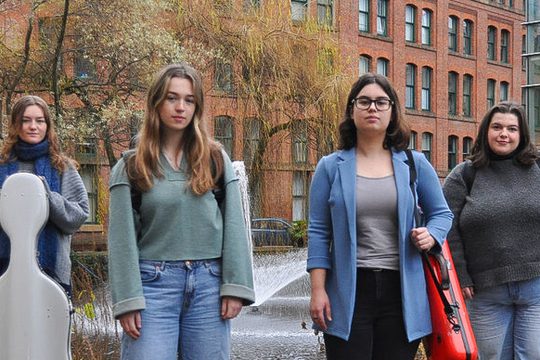 Four women standing holding musical instrument cases in front of a tree.