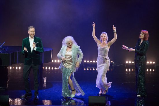Three members dancing in sparkly dresses while a man in a green velvet suit claps his hands