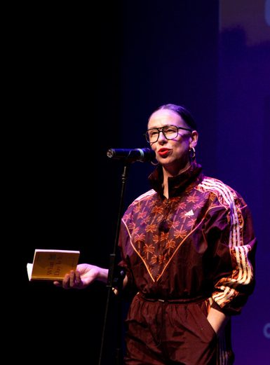 woman with glasses and red lipstick standing at a microphone, wearing a maroon tracksuit with a book in her hand