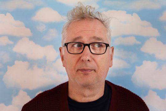 Henry with glasses annd black shirt stand face on in front of a background of clouds looing to the side