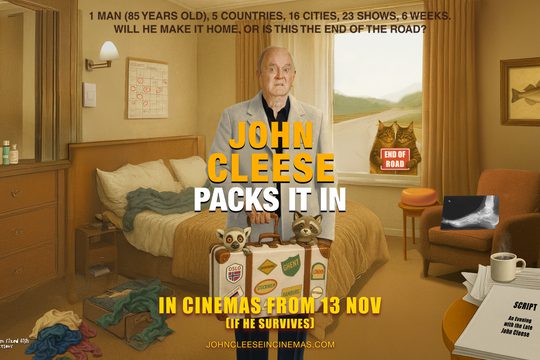 John Cleese in a white suit jacket holding a suitcase with stickers on it.