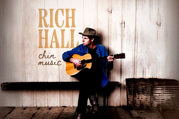 Rich Hall sitting on a bench in a blue shirt, playing guitar