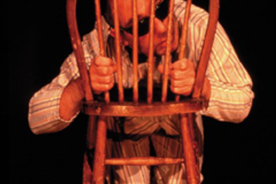 Man in pyjamas and sunglasses peeping through the slats on the back of a wooden chair