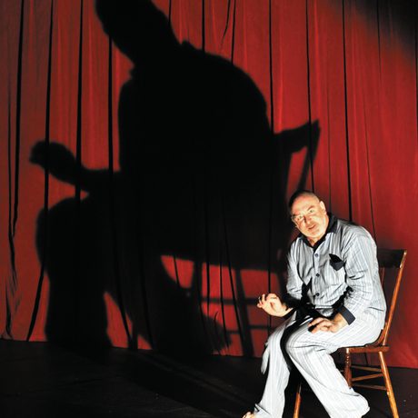 Man in pyjamas sat on a chair with a large looming shadow cast on the red curtain behind him