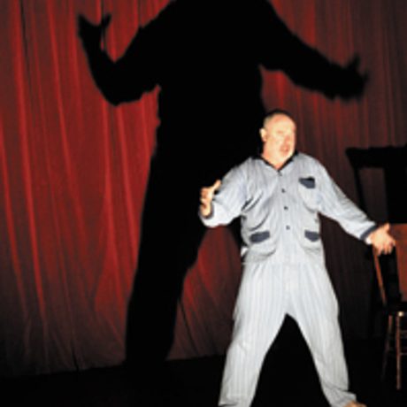 Man in pyjamas, stood up, arms out stretched, with large looming shadow cast on the red curtain behind
