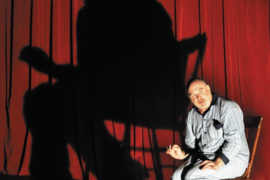 Man in pyjamas sat on a chair with a large looming shadow cast on the red curtain behind him